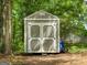 Charming gray and white storage shed in wooded backyard features double doors and adjacent yard tools at 45 Plantation Ln, Locust Grove, GA 30248