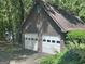 A two-car garage with white doors and brown siding is surrounded by mature trees and shrubbery at 1627 Leguin Mill Rd, Locust Grove, GA 30248