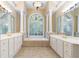 Bright main bathroom featuring dual sinks, white cabinetry, decorative mirrors, and a large tub beneath an arched window at 4567 Brigade Ne Ct, Roswell, GA 30075