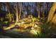 A stone retaining wall with a waterfall feature, shed, pond and surrounding forest at dusk with landscape lighting at 7265 Hunters Branch Dr, Sandy Springs, GA 30328
