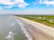 Wide aerial view of a beach with waves lapping onto the shore, showcasing nearby homes and blue skies at 3301 Palm Blvd, Isle of Palms, SC 29451