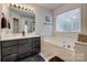 A bright bathroom featuring a soaking tub under a window, a glass-enclosed shower, and a dark vanity with double sinks at 1140 Landsdown Dr, Salisbury, NC 28147