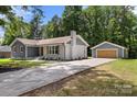 This cozy home has gray siding, brick chimney, and a detached garage with a long driveway, surrounded by lush greenery at 7141 Pitchfork Ln, Mint Hill, NC 28227