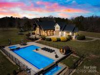 A dusk aerial view of a backyard pool area, including a hot tub, sauna, lounge chairs, and waterfall feature at 12613 Oak Grove Rd, Stanfield, NC 28163