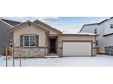 Tan house with stone accents, a two-car garage, and landscaping; winter scene at 144 Wooten Ave, Brighton, CO 80601