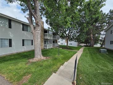 Low-angle exterior of apartments with lush green lawns, mature trees, and gray siding accented by stone chimney and dark shutters at 14433 E Jewell Ave # 101, Aurora, CO 80012