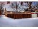 Snowy view of a brick home's exterior featuring a wooden fence, garage, and mature trees in a winter landscape at 1817 E Center Ave, Denver, CO 80209