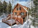 Beautiful view of log cabin with a spacious wooden deck and stairs surrounded by mature trees in a serene, snow-covered setting at 11498 Pauls Ln, Conifer, CO 80433