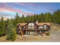 Beautiful rustic log home featuring large windows, a stone chimney, wood deck, and mature trees at 25952 Fern Gulch Rd, Evergreen, CO 80439