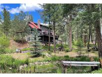 Stunning multi-story home with stone accents and a red metal roof surrounded by lush green trees and a tranquil pond at 9716 Canon Dr, Sedalia, CO 80135