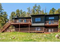 Two-story home with a large deck, wood siding, chimney, and stairs against a backdrop of trees and blue sky at 34075 Grouse Ln, Evergreen, CO 80439