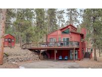 Red two-story home with a large deck, an overhang with blue chairs, surrounded by tall evergreen trees and a small shed at 6997 S Columbine Rd, Evergreen, CO 80439