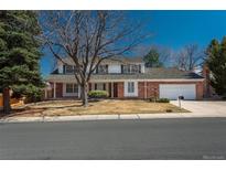 Traditional brick home featuring a two-car garage, manicured lawn, mature trees, and blue sky at 3926 S Magnolia Way, Denver, CO 80237