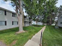 Low-angle exterior of apartments with lush green lawns, mature trees, and gray siding accented by stone chimney and dark shutters at 14433 E Jewell Ave # 101, Aurora, CO 80012