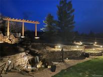 Inviting backyard with stone terraced landscaping, waterfall feature, wooden pergola, and landscape lighting at twilight at 580 Summer Mist Cir, Castle Rock, CO 80104