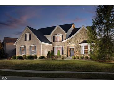 Elegant brick and stone home featuring a manicured lawn, lush landscaping, and a welcoming arched entryway at dusk at 14707 Thor Run Dr, Fishers, IN 46040