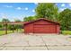 View of a red garage featuring a carport and a cracked concrete driveway, set against a lush green backdrop at 6123 W 46Th St, Indianapolis, IN 46254