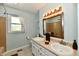 Bathroom with a double sink vanity, soft blue walls, a window with floral curtains, and a tiled shower at 8548 Scarsdale W Dr, Indianapolis, IN 46256