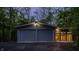 Evening view of the modern garage with two doors, accent lighting, and a separate entrance, all set in a wooded area at 12469 Springmill Rd, Carmel, IN 46032