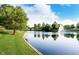 Scenic community pond with fountain, green grass, and mature trees against a partly cloudy sky at 2436 Gwinnett St, Carmel, IN 46032