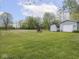 Expansive grassy backyard features a shed and a garage, with a brick fire pit, bordered by lush trees under a blue sky at 4305 Clinton St, Lawrence, IN 46226