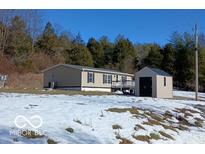 Tan mobile home with deck and storage shed, set on a snowy lot with wooded backdrop at 12160 W Baker Hollow Rd, Columbus, IN 47201