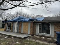 Damaged brick home with tarp on roof, boarded window, and debris strewn about at 3514 Luewan Ct, Indianapolis, IN 46235