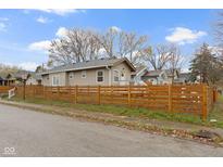 Tan home featuring a wood fence, well-maintained lawn, and tree line, all showcased under a bright, partly cloudy sky at 802 N Tuxedo St, Indianapolis, IN 46201