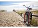 A bicycle rests on a wooden railing near a sandy path leading to a beach under a bright, sunny sky at 1609 Seachase Way, North Myrtle Beach, SC 29582