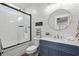 Bathroom featuring blue vanity, round mirror, and tub-shower with glass doors and white subway tile at 5100 Ocean Blvd. N, North Myrtle Beach, SC 29582