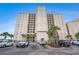 Condo building's exterior with a parking area, palm trees, and a blue sky backdrop at 2151 Bridge Ct. # 1-305, North Myrtle Beach, SC 29582