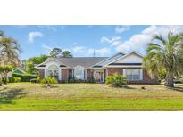 Charming one-story home featuring a well-manicured lawn, brick facade, and mature palm trees, with blue skies in the background at 4104 Friendfield Trace, Little River, SC 29566