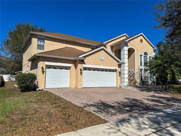 Two-story tan home featuring a three-car garage, brick driveway, and a well-manicured lawn under a clear blue sky at 2387 Laurel Blossom Cir, Ocoee, FL 34761