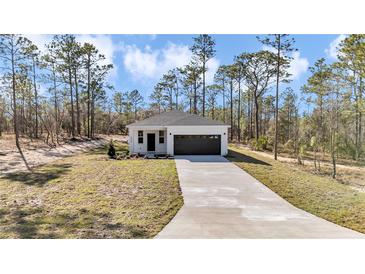 Charming single-story home featuring a white exterior, dark garage door, and a well-manicured lawn surrounded by tall trees at 406 Nw Narcissus Rd, Dunnellon, FL 34431