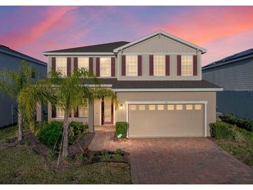 Two-story home featuring tan siding, red shutters, a brick driveway and an attached two-car garage at 613 Bloom Ter, Davenport, FL 33837