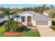 Beautiful single-story home featuring a gray roof, a three-car garage, and lush landscaping with vibrant plants and palm trees at 27023 Old English Ct, Leesburg, FL 34748