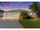 Beautiful home featuring a three-car garage, light-blue exterior, well-manicured lawn, and decorative driveway at dusk at 4878 Chitty Chatty Run, The Villages, FL 32163
