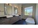 Modern bathroom with dual sinks, gray cabinets, black countertop, a free-standing tub, and a large window at 16902 Vinci Way, Montverde, FL 34756