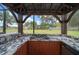 Outdoor kitchen with granite countertop, gas stove and wooden cabinets under a covered structure, surrounded by lush greenery at 3934 Se 110Th St, Belleview, FL 34420