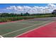 Outdoor tennis court featuring red and green surface, surrounded by fencing and lush greenery under a sunny, cloudy sky at 215 Grovepark Dr, Davenport, FL 33837