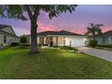 Dusk view of single-story home featuring a two-car garage, manicured lawn and mature trees under a colorful sky at 467 Weston Manor Dr, The Villages, FL 32162