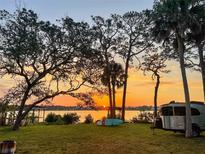Scenic waterfront property featuring a vibrant sunset, lush green lawn, and mature shade trees at 1949 Bayview Dr, New Smyrna Beach, FL 32168
