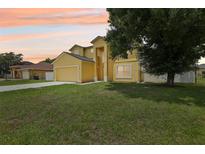 Two-story home with yellow stucco, attached garage, manicured lawn and mature trees at sunset at 670 Bluebill Ct, Poinciana, FL 34759