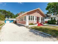Charming pink home with red steps and a gravel driveway, surrounded by lush greenery under a clear blue sky at 205 Mary Ave, New Smyrna Beach, FL 32168