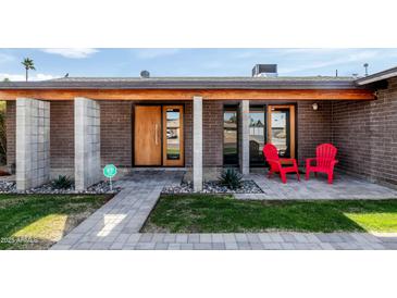 Charming brick home featuring a stylish wood door, modern concrete accents, and vibrant red chairs on the front porch at 3519 E De Poe Ln, Phoenix, AZ 85028