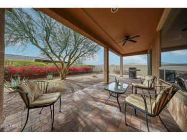 Relaxing covered patio featuring brick flooring, seating, and view of the landscaped backyard with blooming flowers at 17555 W Glenhaven Dr, Goodyear, AZ 85338