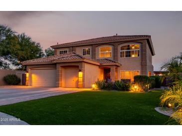Inviting two-story home featuring lush green grass, a three car garage, and warm exterior lighting at twilight at 9569 W Redbird Rd, Peoria, AZ 85383