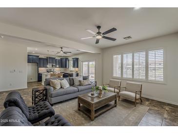 Comfortable living room with a grey sofa, ceiling fan, kitchen access, and bright natural light at 40759 W Bravo Dr, Maricopa, AZ 85138