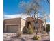 Tan home featuring a two-car garage, desert landscaping, and a decorative tree in the front yard under a partially cloudy blue sky at 5705 S Pinnacle Ln, Gold Canyon, AZ 85118