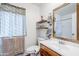 Bright bathroom featuring a white vanity, wood-framed mirror, open shelves, and a window with floral curtains at 10407 W Mission Ln, Sun City, AZ 85351